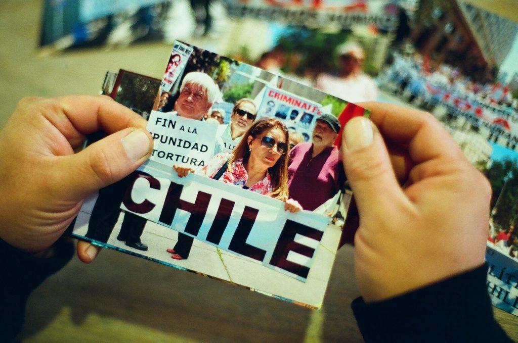 Manos sosteniendo revista o periódico con palabra CHILE visible y fotografías de personas en portada, sobre mapa