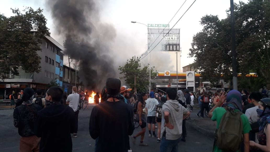 "Entre Cumming y Brasil": La fundación del nuevo Chile Multitud en calle durante protesta con edificio en llamas de fondo y humo denso en el cielo