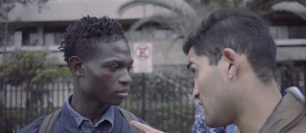 Dos hombres jóvenes frente a frente en conversación intensa, con viviendas y vegetación de fondo