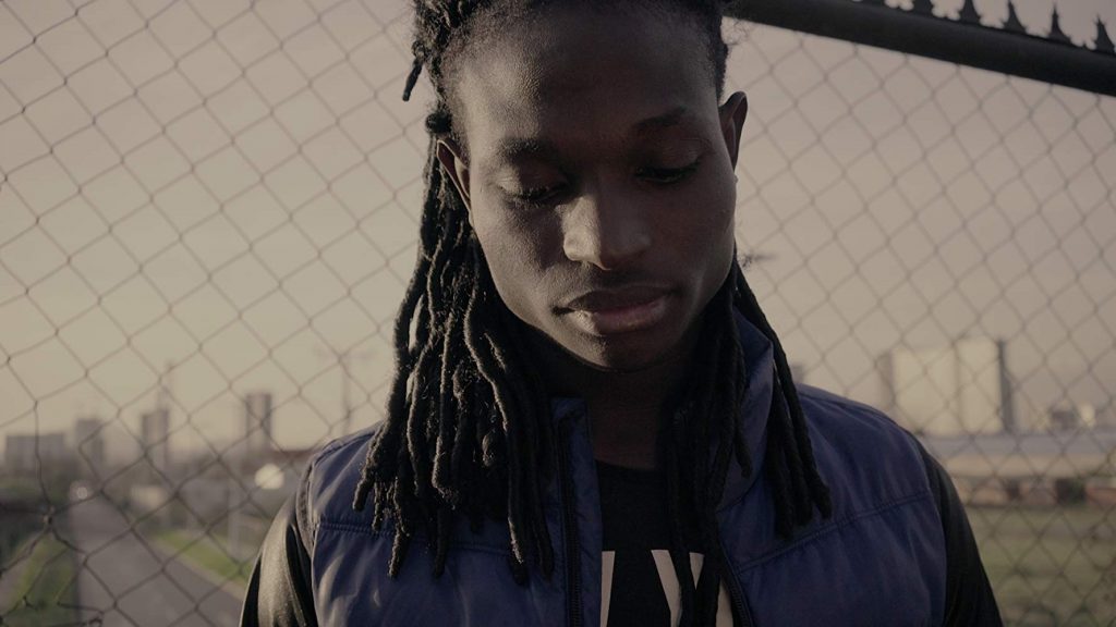 Joven con rastas mira hacia abajo en una azotea urbana con ciudad al fondo, ambiente cinematográfico