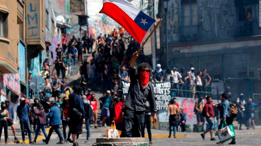 Multitud de manifestantes en plaza urbana con bandera de Chile roja y azul ondeando, protestando contra dictadura