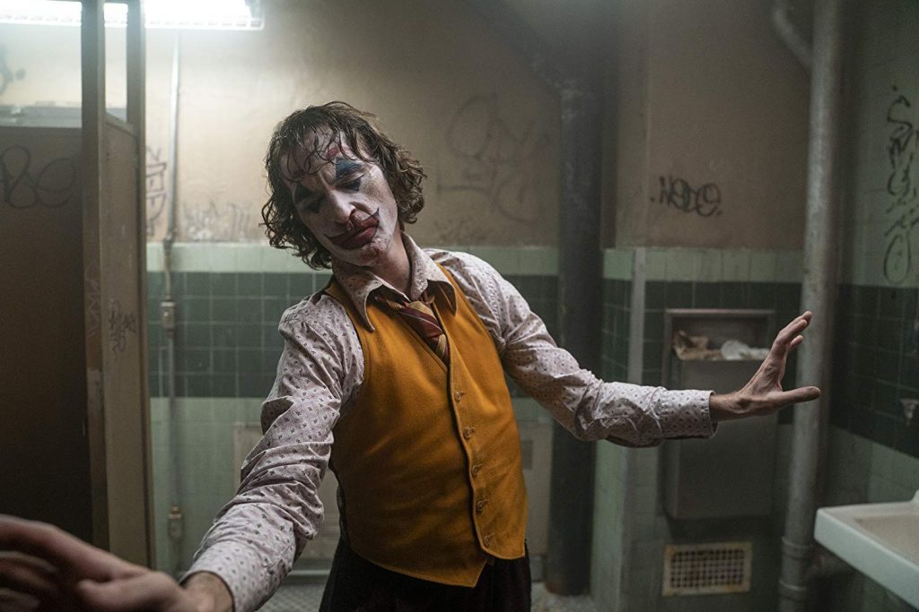 Joaquin Phoenix como Joker en un cuarto de baño, gesticulando con expresión desquiciada y ropa desgastada