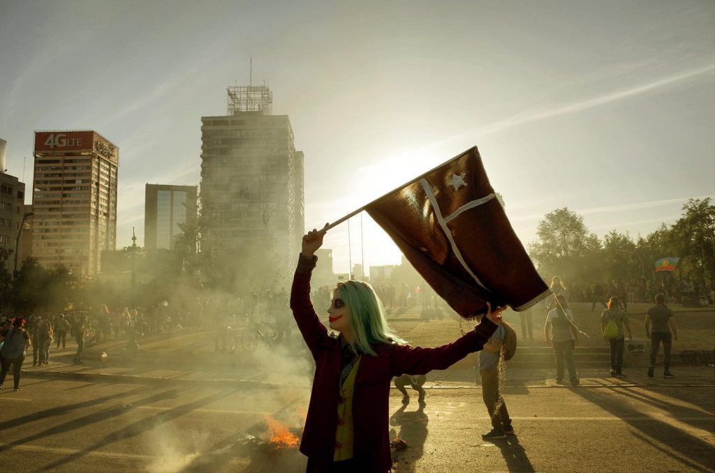 "Terremotos y miedos": Una retrospectiva al levantamiento popular de 2019 Persona sostiene bandera durante manifestación en calle urbana con edificios y humo de fondo