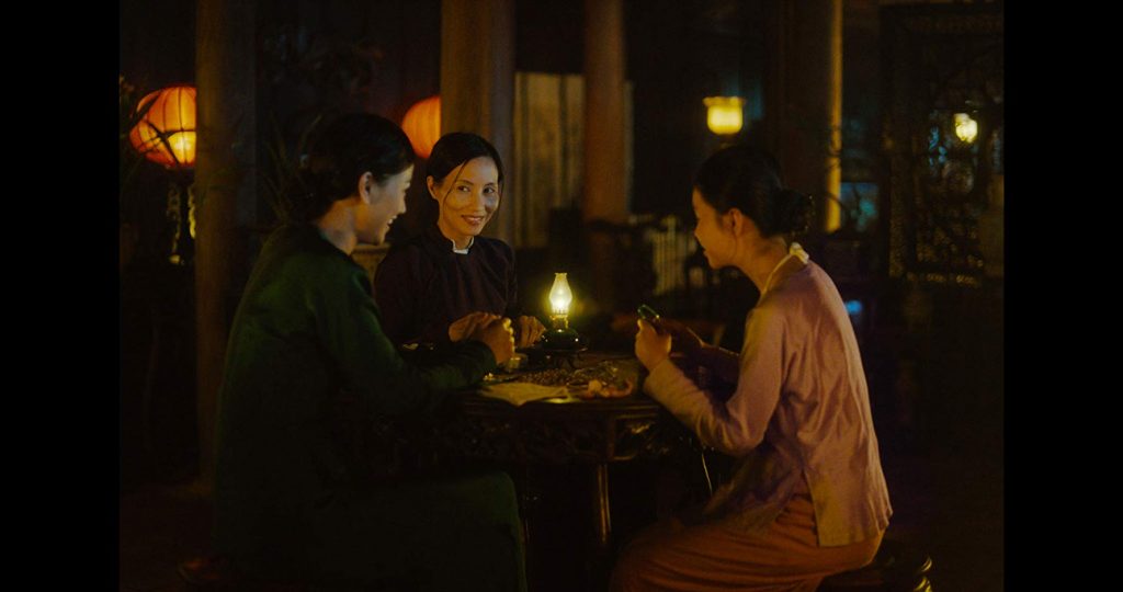 “La tercera esposa”: Una historia de mujeres Escena nocturna de diálogo entre una mujer y varios hombres alrededor de una mesa iluminada por velas en ambiente