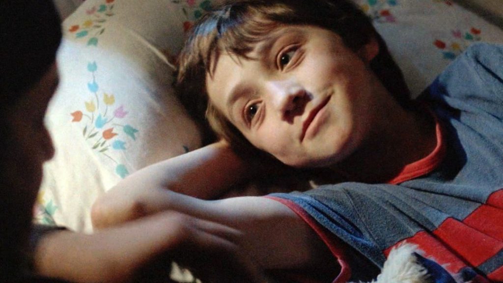 Niño acostado en cama con almohada floral, mirando hacia la cámara en escena del film Decálogo de Kieślowski