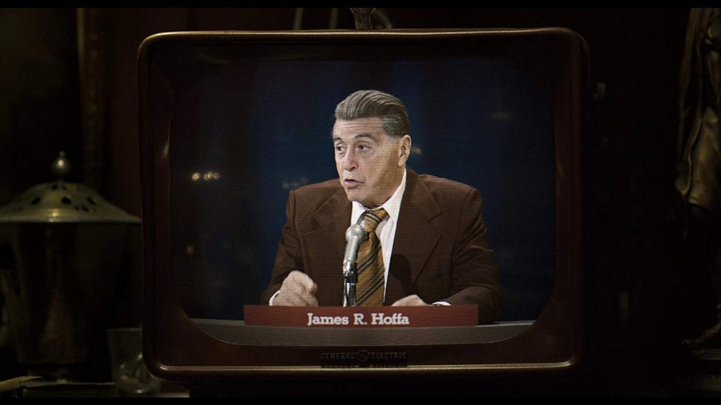 Hombre mayor con gafas en traje marrón sentado ante escritorio, iluminado en estudio de televisión retro