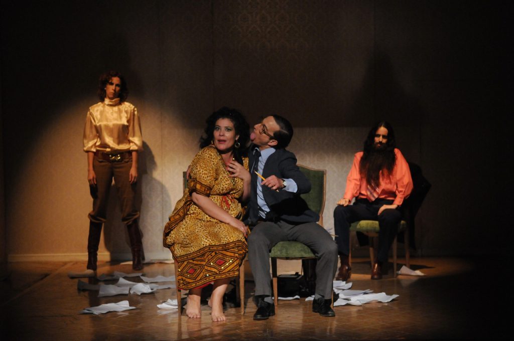 Cinco actores en escena bajo iluminación teatral: cuatro de pie y uno sentado, en representación de la obra