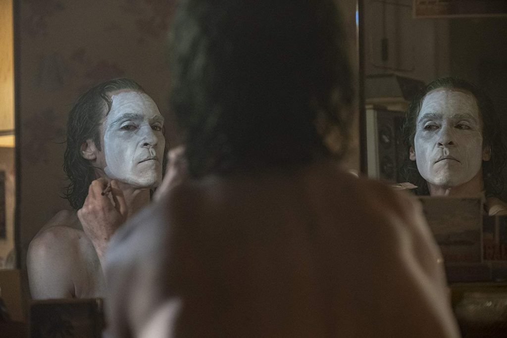 Hombre de mediana edad observándose en espejo con expresión perturbada y maquillaje de payaso blanco en rostro