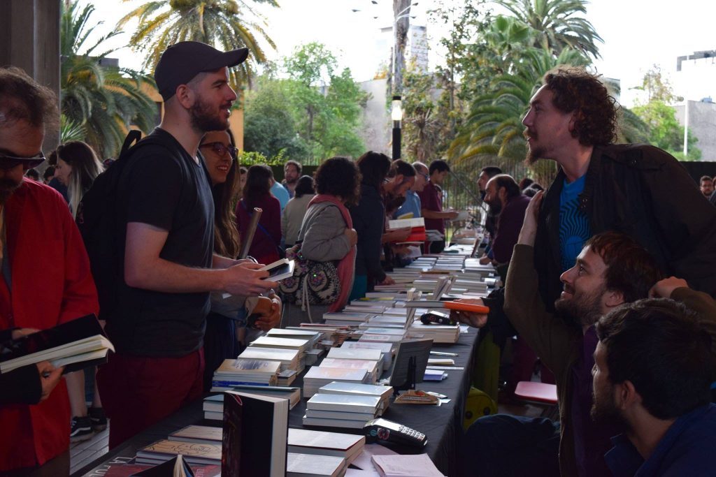 Personas en una feria de libros al aire libre con mesas de venta, árboles y palmeras de fondo en ambiente soleado