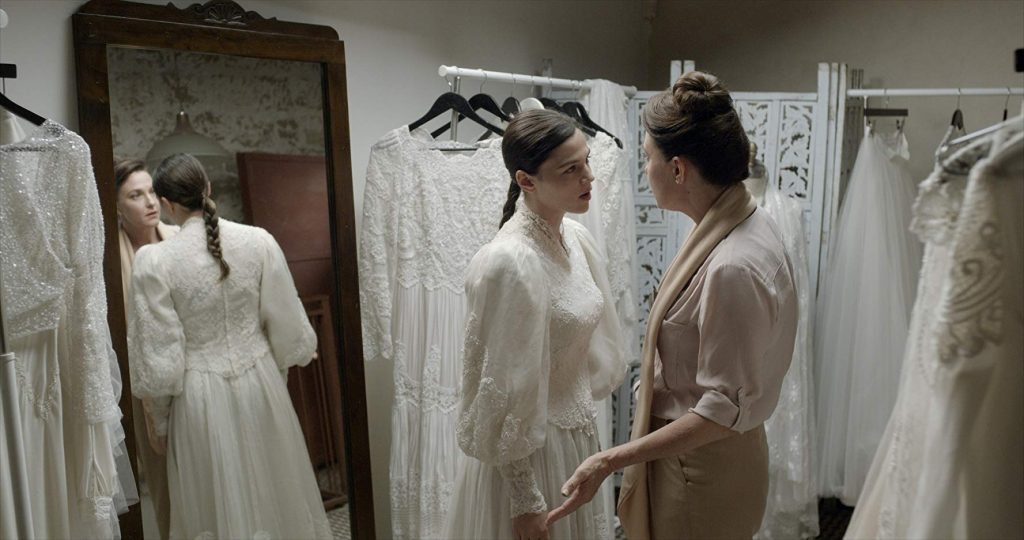 Taller de confección de vestidos de novia, costurera ajustando un traje blanco mientras cliente observa en espejo