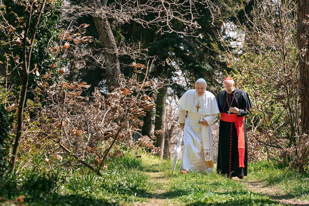 Dos hombres vestidos con túnicas religiosas caminan por un sendero arbolado en ambiente de bosque