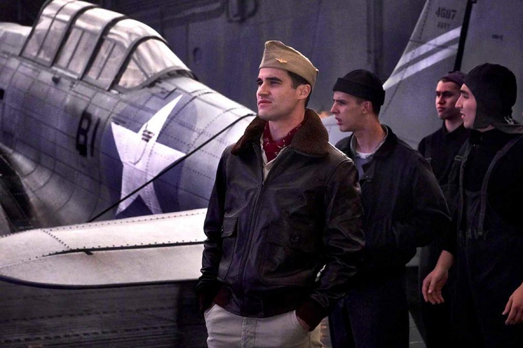 Hombre con gafas observa maqueta de avión militar junto a otras personas en set de producción cinematográfica