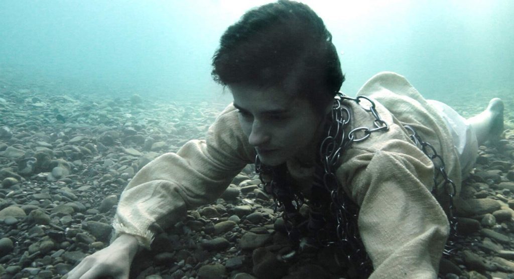 Hombre bajo el agua en posición de buceo, mirando hacia la cámara con expresión contemplativa en ambiente submarino