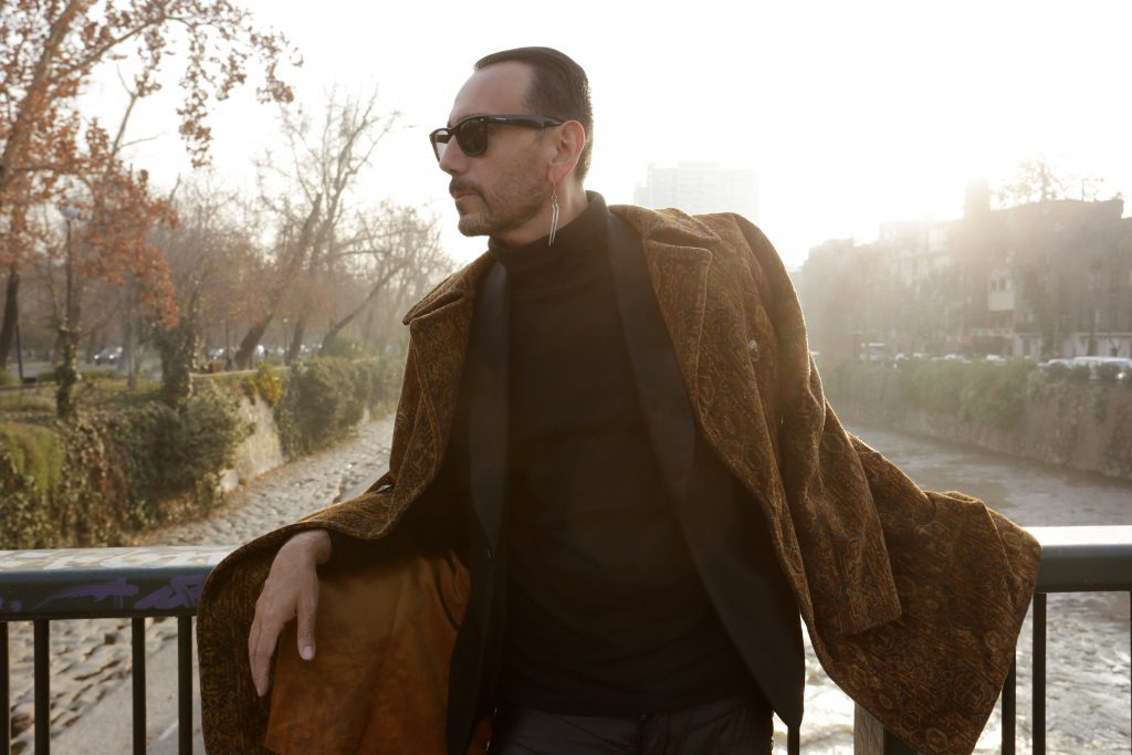 Hombre de mediana edad con gafas oscuras y abrigo marrón, apoyado en una barandilla junto a un canal arbolado