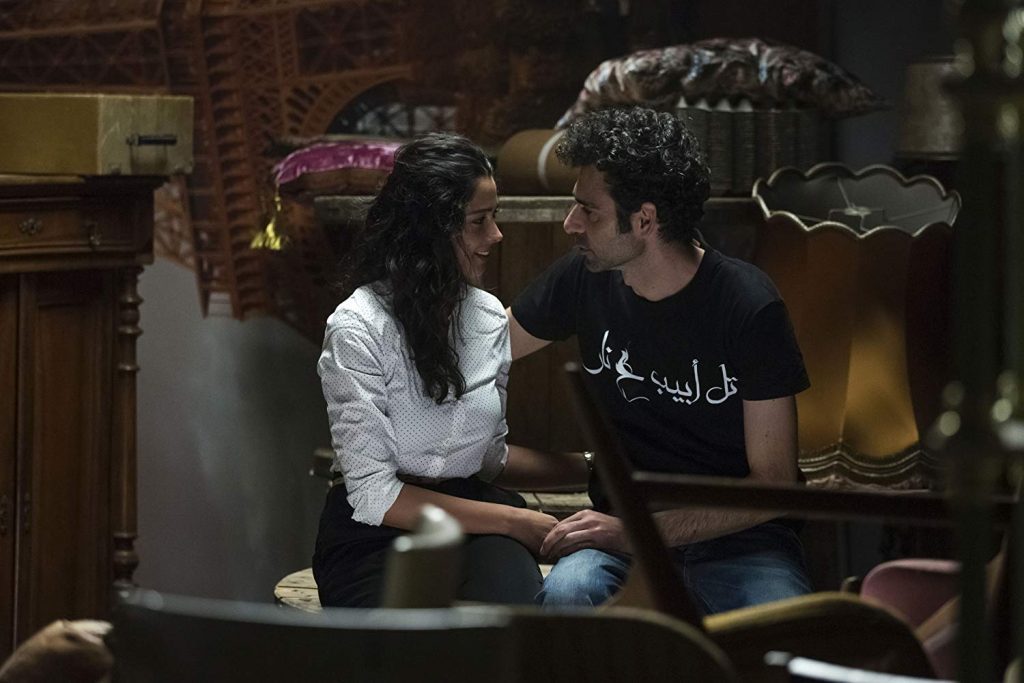 Dos personas conversan en un interior oscuro y cálido. Hombre de cabello oscuro y mujer con cabello rizado se miran