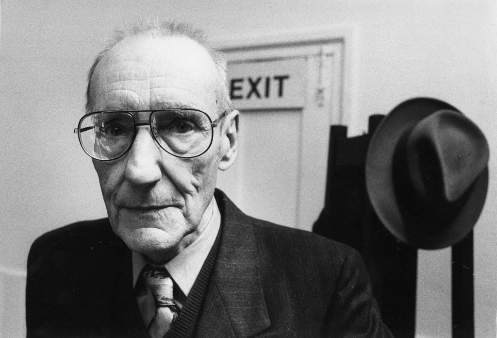 William Burroughs con gafas y traje oscuro en Londres, 1988, junto a un cartel de salida