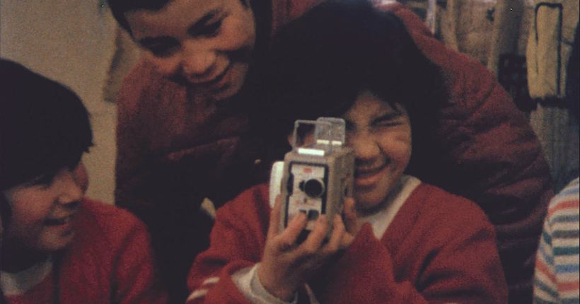 Grupo de niños en ropa roja de los años 70, uno sostiene una cámara fotográfica mientras sonríe a la cámara