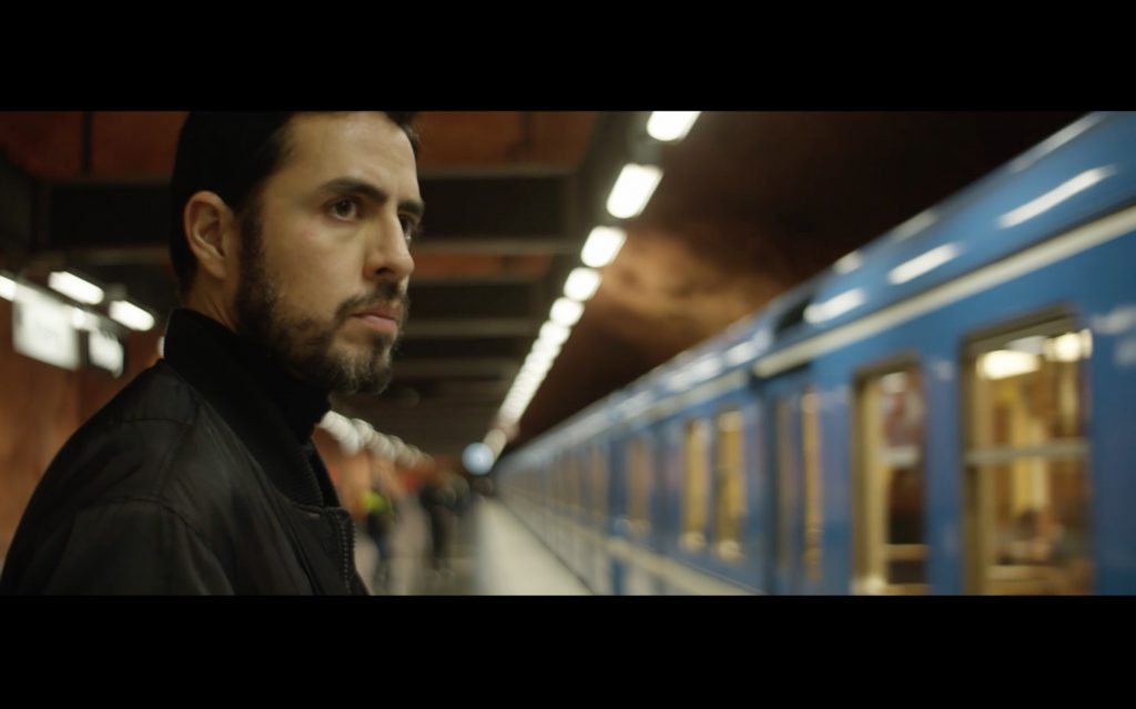 "El estado imaginario": El filme cuyo guión se redactó con la colaboración de la escritora Andrea Jeftanovic Hombre de barba con chaqueta negra en andén de estación de tren azul, mirando hacia cámara con expresión seria