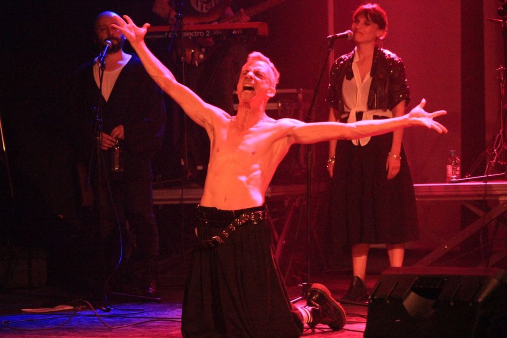 Actor sin camisa en escena teatral iluminada en rojo, con brazos extendidos, acompañado por músicos al fondo