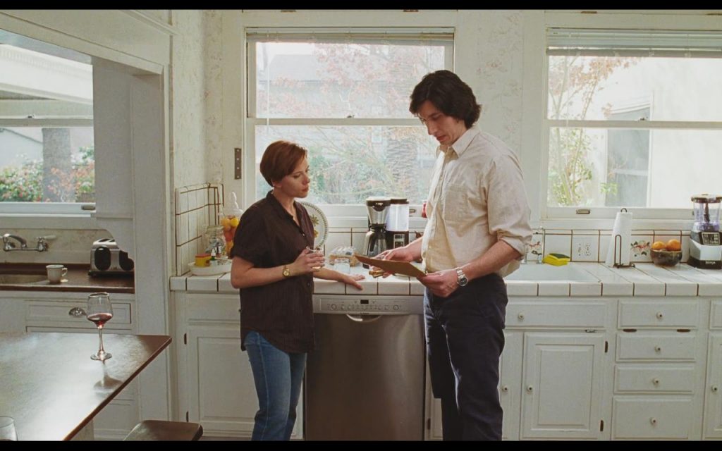 Dos hombres en una cocina moderna intercambian una caja; uno viste camisa marrón y el otro camisa beige clara