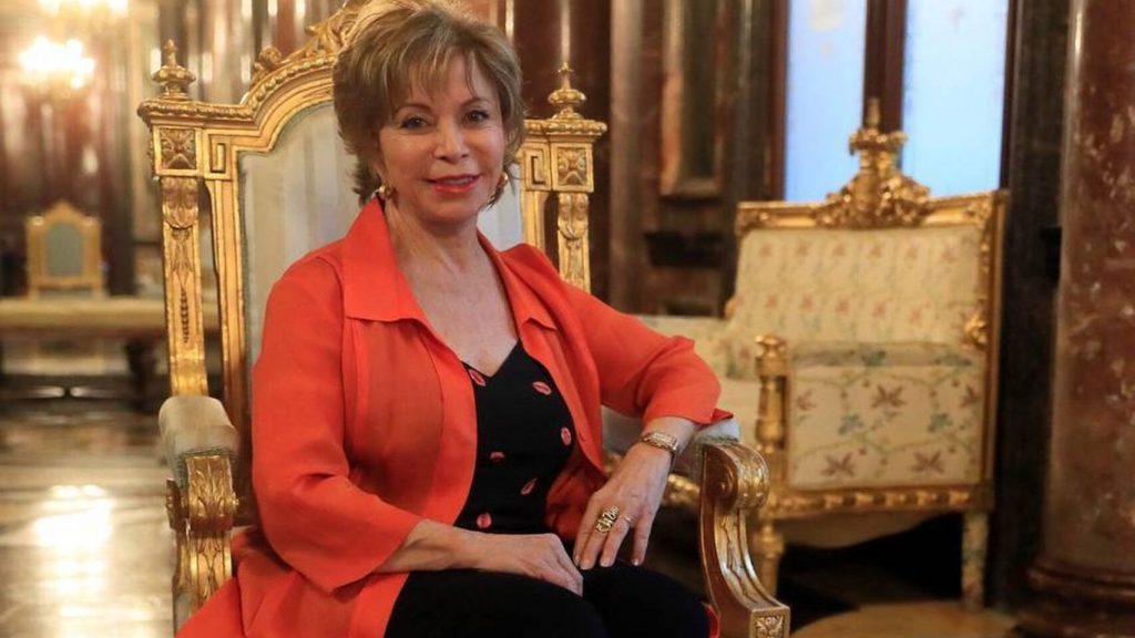 "Escritores célebres, conocidos y... los otros": El club de la pelea Isabel Allende Llona, escritora chilena, sentada en sillón dorado con blazer rojo coral sobre fondo de salón elegante