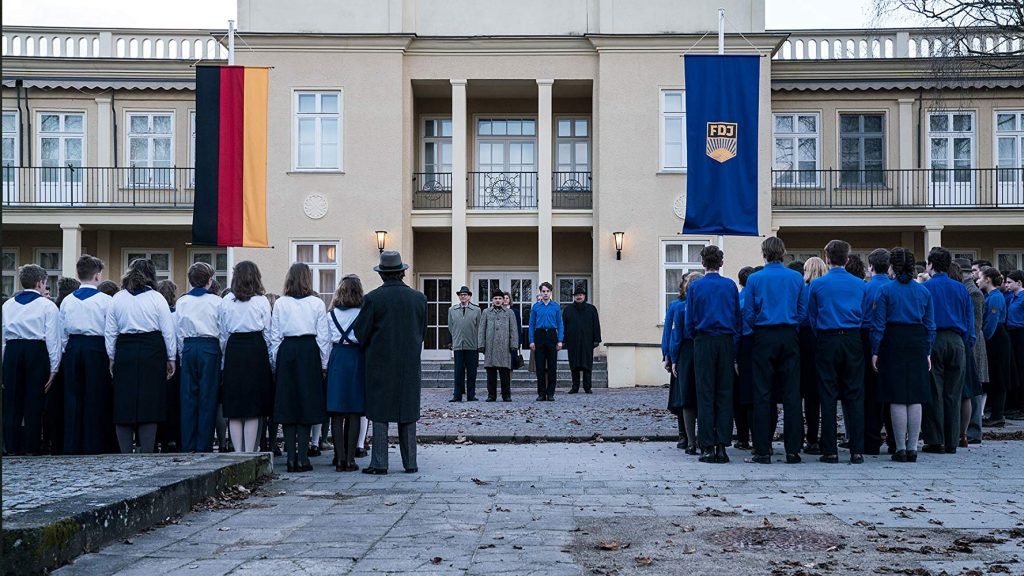 "La revolución silenciosa": Una historia de traiciones y engaños detrás de la "Cortina de Hierro" Dos grupos de personas en uniforme escolar enfrentadas en patio de edificio histórico con fachada de colores pastel