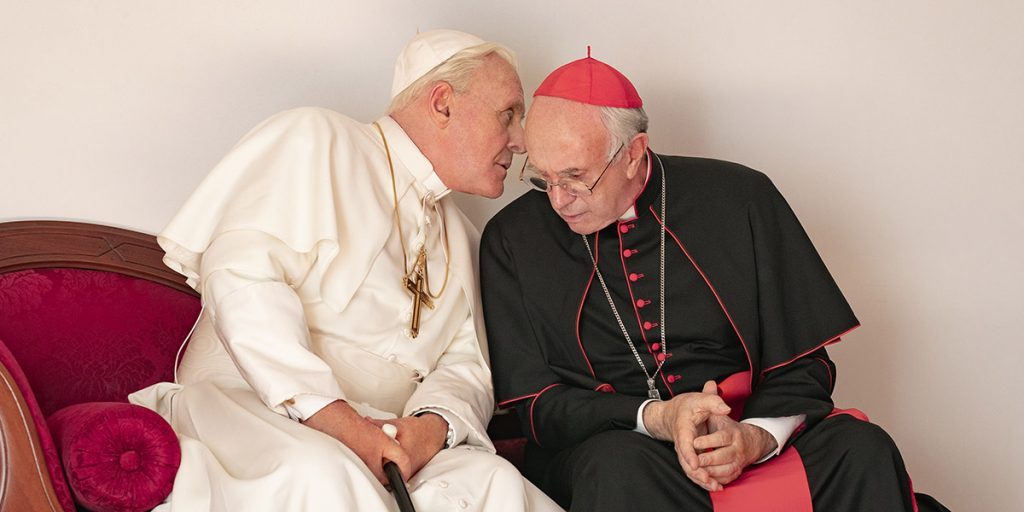 Dos eclesiásticos en conversación íntima sentados en sofá de terciopelo rojo, uno de blanco y otro de negro con cruz