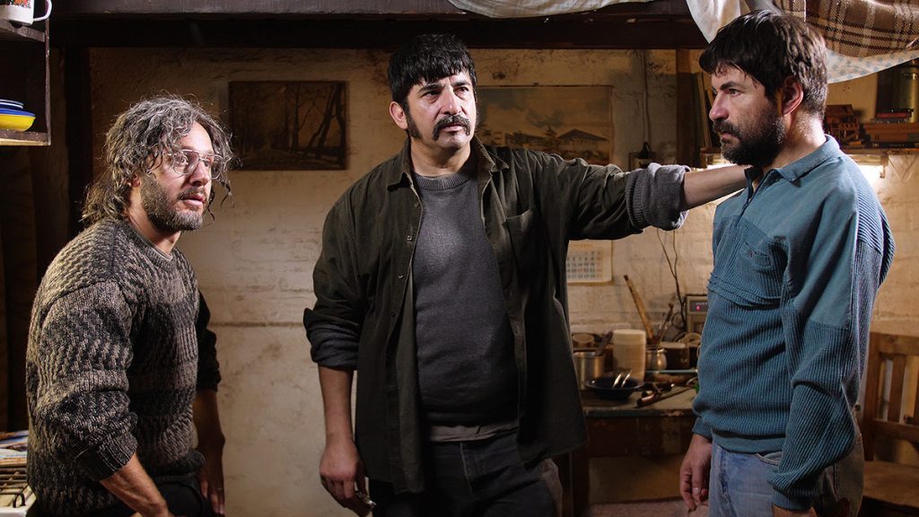 Tres hombres en conversación dentro de un espacio de madera rústica, uno gesticula mientras los otros lo observan