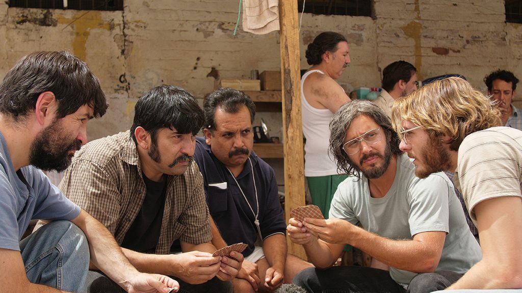 "Pacto de fuga": La valentía en un Chile de miedos Grupo de hombres en círculo dentro de un espacio austero, conversando intensamente durante lo que parece ser un ensayo