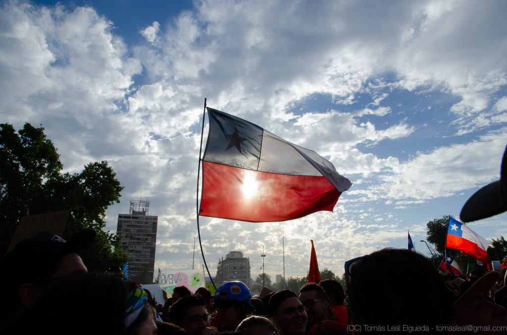 Bandera de Chile ondeando al atardecer durante manifestación callejera con multitud de personas de fondo