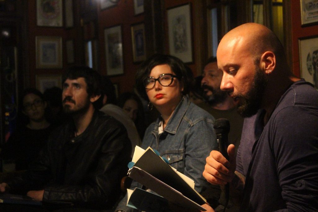 Tres hombres en interior con paredes de cuadros. El del centro sostiene un libro. Ambiente de presentación o evento