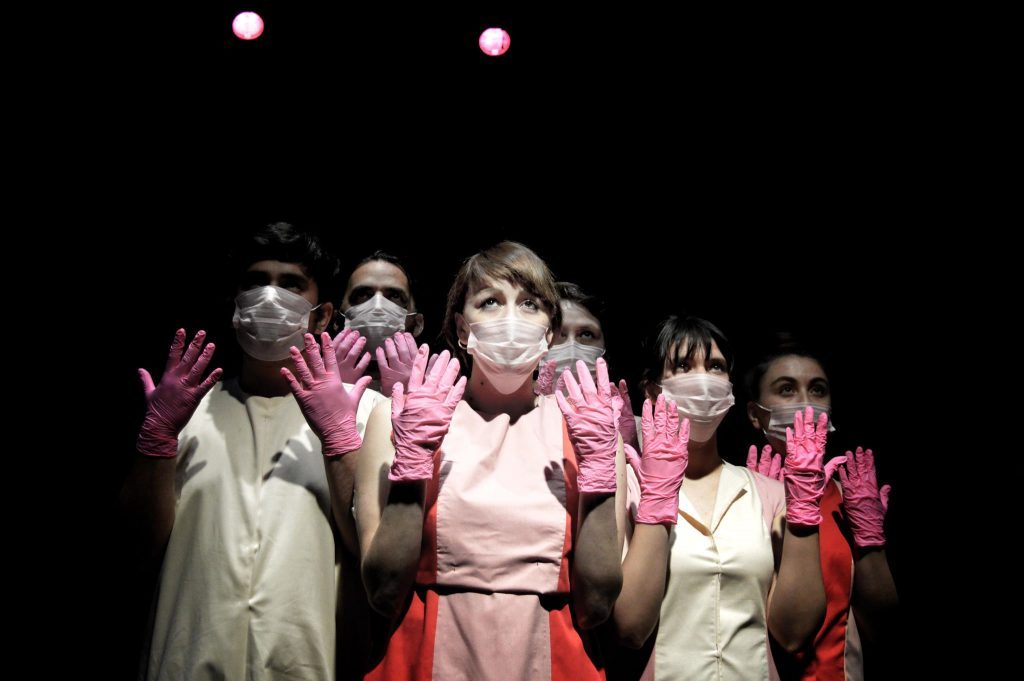 Actriz en escena de teatro con traje rosa claro rodeada de otros actores en vestuario vintage bajo luces de escena