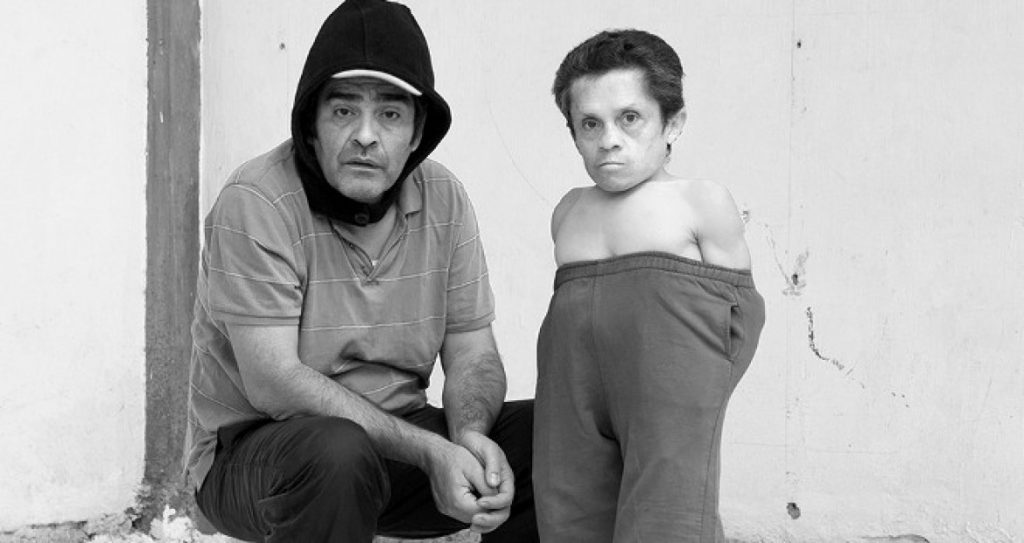 Dos hombres sentados contra una pared blanca en fotografía en blanco y negro, uno con gorro y otro de perfil