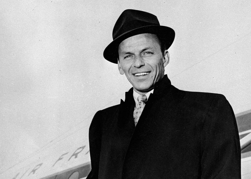Frank Sinatra sonriendo, vistiendo traje oscuro y sombrero fedora negro, fotografía en blanco y negro