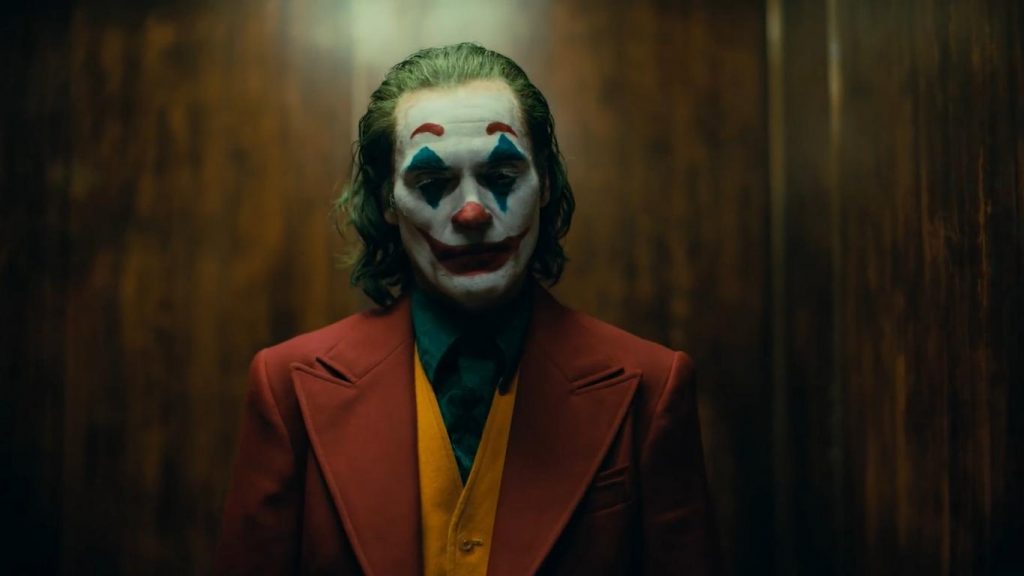 Hombre con maquillaje de payaso blanco, nariz roja y sonrisa exagerada, vistiendo traje rojo y camisa amarilla