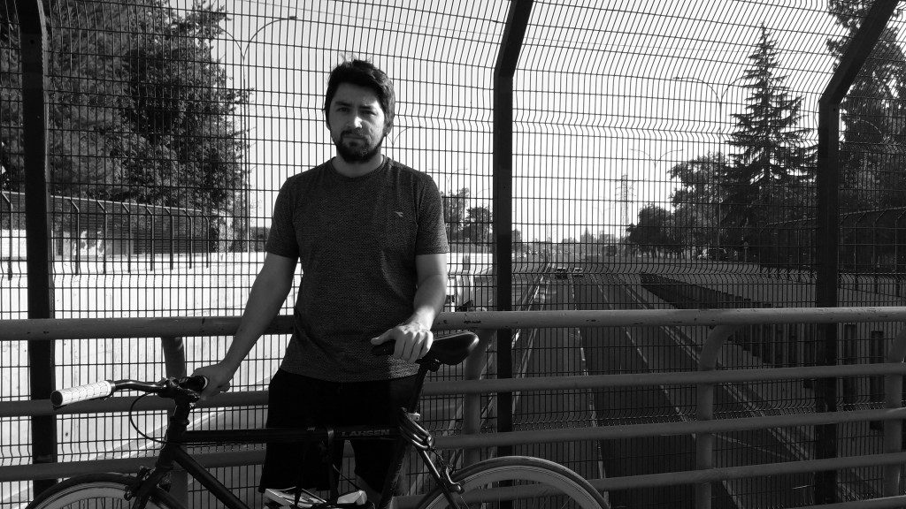 Nicolás Meneses en blanco y negro, de pie junto a una bicicleta tras una valla metálica en un parque