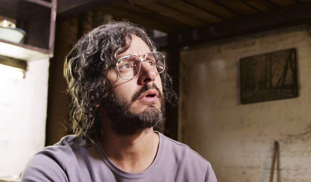 "Pacto de fuga": La emoción artística de una sorpresa Hombre con cabello largo y gafas, expresión contemplativa, en interior con paredes de hormigón y cuadro al fondo
