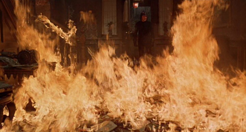 Intenso incendio con llamas naranjas consumiendo libros y objetos en una habitación oscura, escena de destrucción