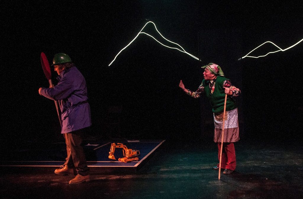 Dos actores en escena teatral nocturna con iluminación azul y roja, uno sostiene una cuerda blanca, junto a objetos