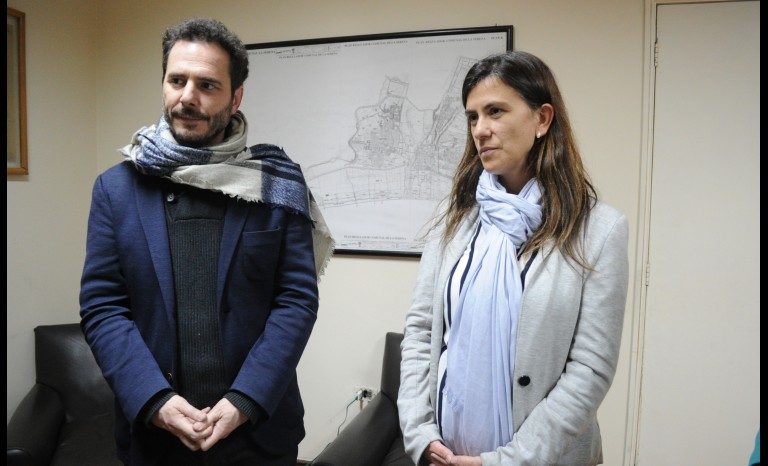 Dos personas en una oficina: hombre con suéter azul y bufanda, mujer con blusa blanca, ambos de pie frente a escritorio