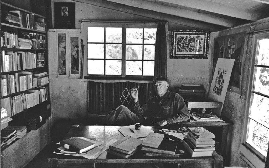 Henry Miller en su escritorio rodeado de libros, papeles y manuscritos en un espacio de trabajo desordenado