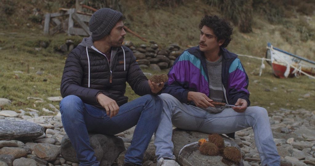 "Los fuertes", de Omar Zúñiga: Secreto en la montaña a la chilena Dos hombres conversan sentados en un tronco en un campamento de montaña con tiendas al fondo