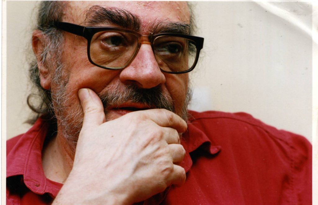 Mario Levrero, escritor uruguayo, retrato en primer plano con gafas oscuras y mano cerca del mentón, fondo neutro