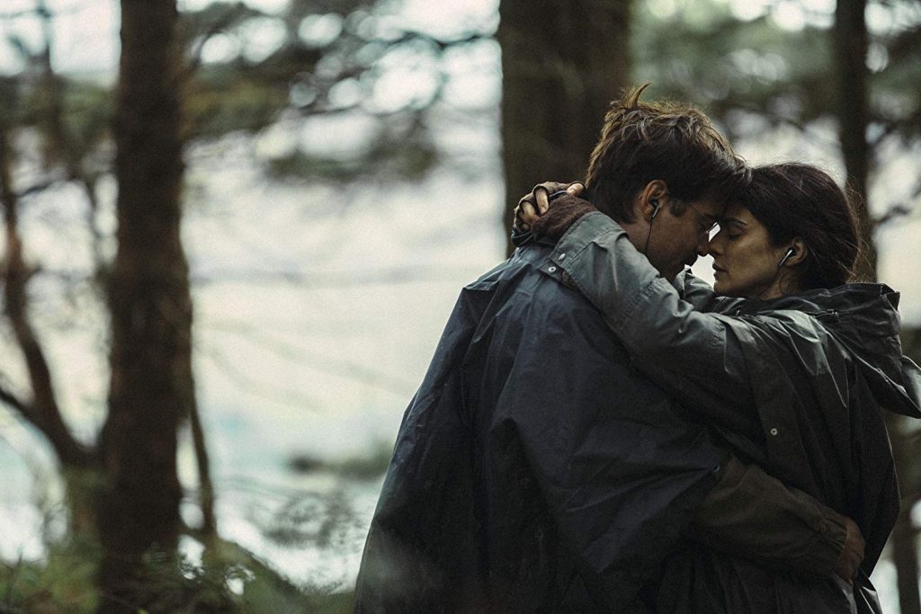 Pareja abraza a un hombre en bosque junto al lago, escena de tensión emocional en ambiente natural sombrío