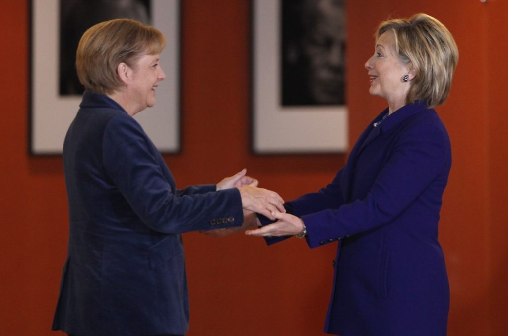 Ángela Merkel e Hillary Clinton se dan la mano en un saludo formal frente a un fondo rojo con marcos en la pared