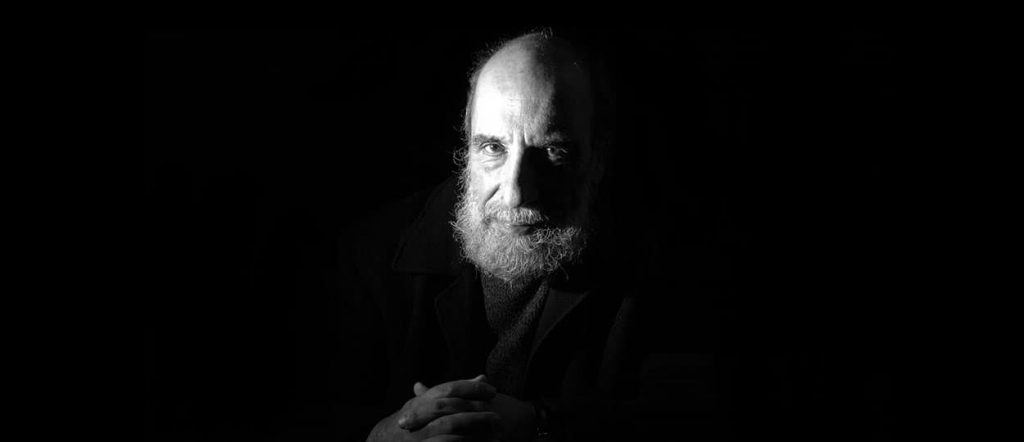Raúl Zurita, poeta chileno, retratado en blanco y negro con barba y expresión contemplativa sobre fondo oscuro