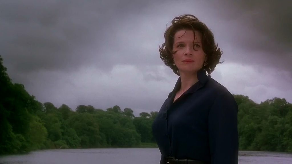 Mujer de cabello castaño ondulado junto a un río bajo un cielo nublado, con expresión serena y contemplativa