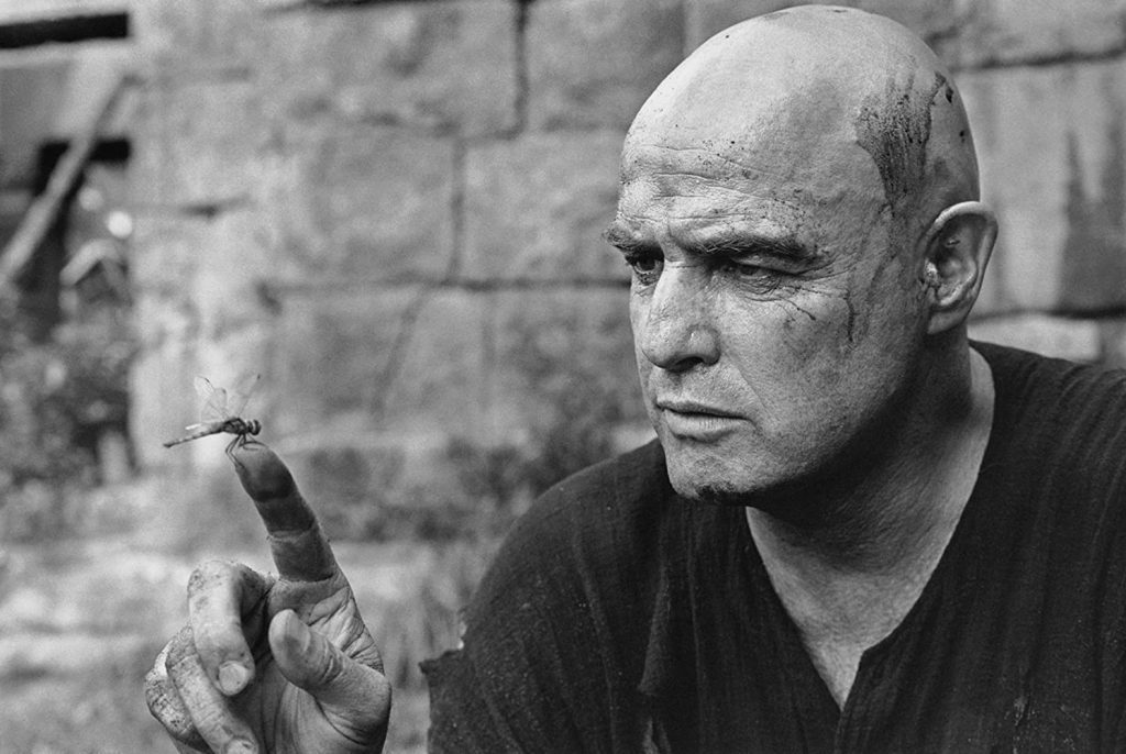 Marlon Brando en primer plano, calvo, fumando, con expresión seria contra pared de ladrillo