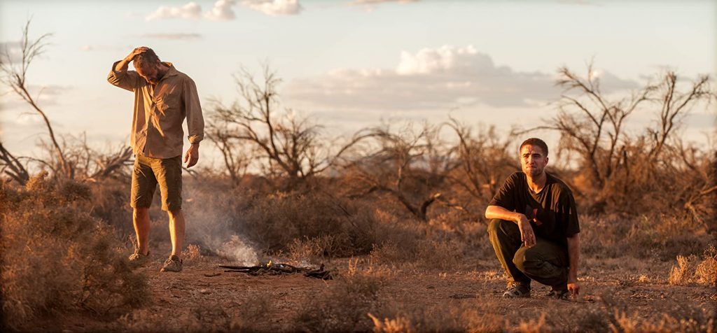 Dos figuras en posición de caza atraviesan un árido paisaje desértico bajo luz dorada del atardecer, con vegetación