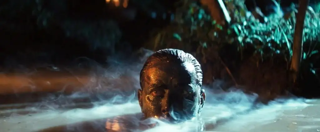 Rostro de hombre cubierto de barro en agua turbia, con vegetación selvática al fondo iluminada por fuego